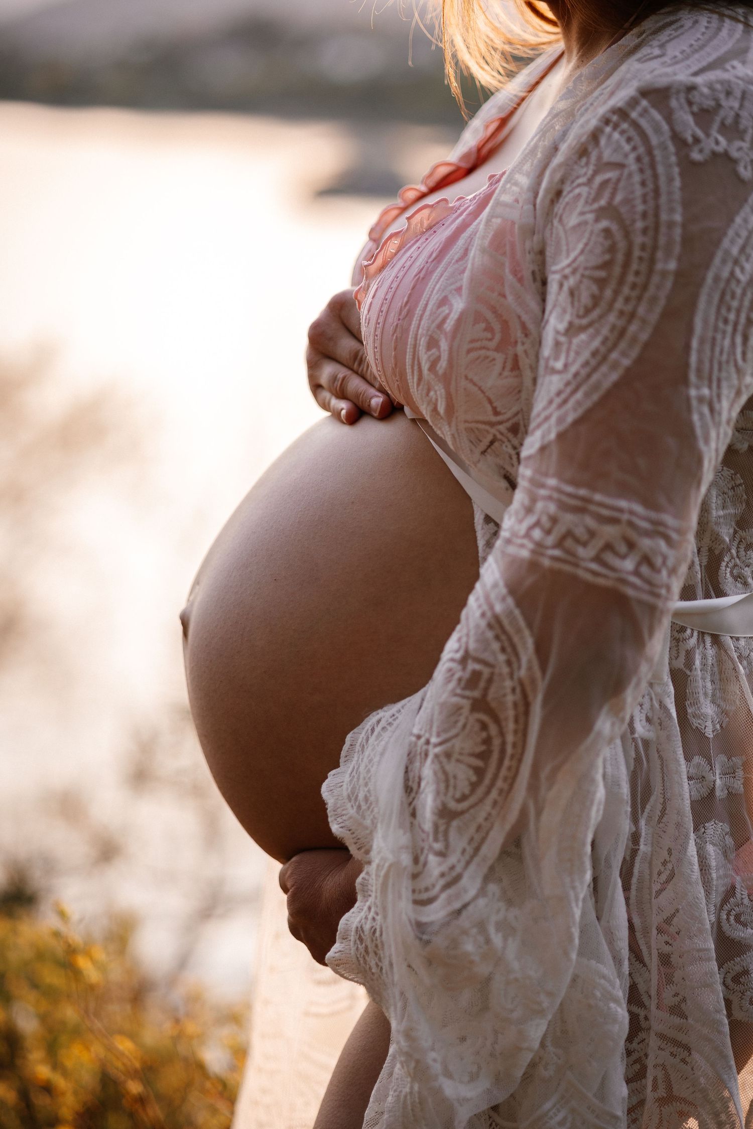 Silhouette of an expecting mother in a lace dress standing by the water during golden sunset.
