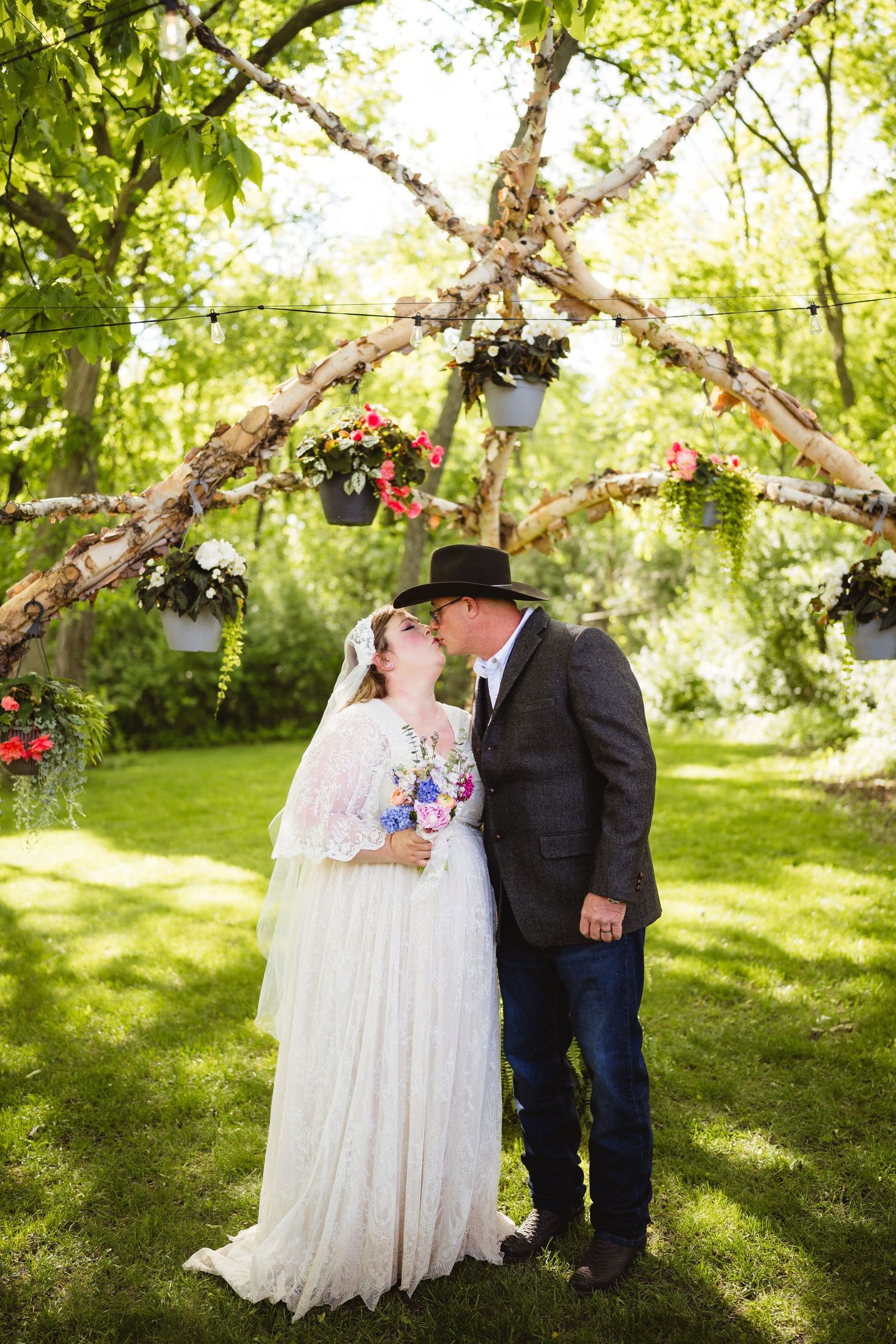 A couple shares a romantic moment under flowering tree branches in a sunlit garden setting.