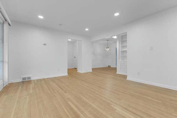 Bright empty living room with light hardwood floors, recessed lighting, built-in shelving, and white walls.