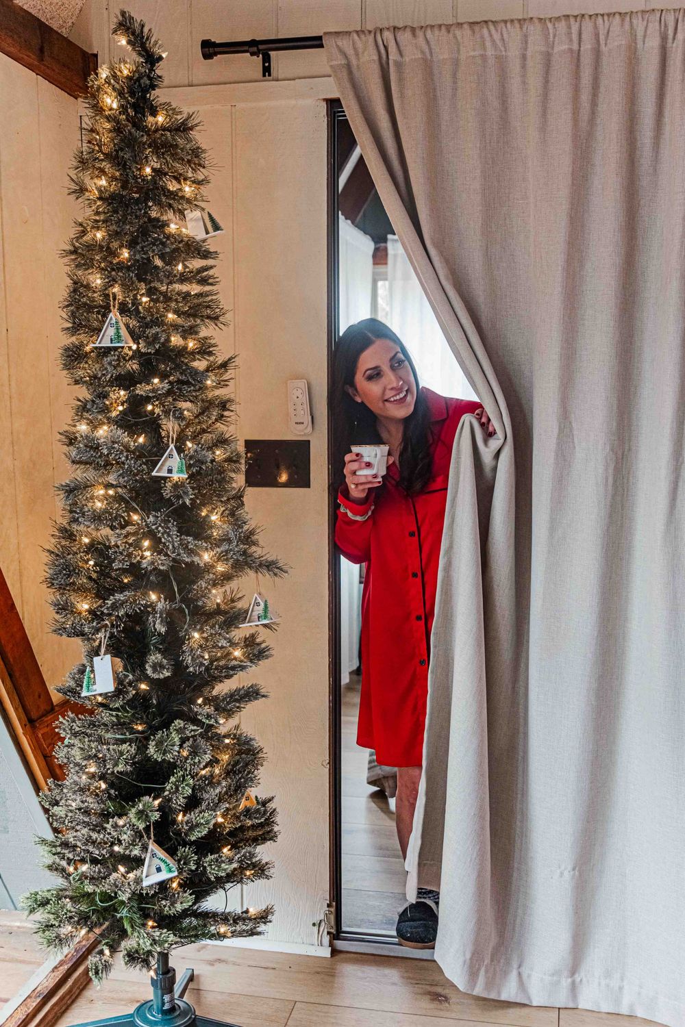 A decorated slim Christmas tree stands next to a curtained doorway where someone peeks through wearing a red dress holding a coffee cup