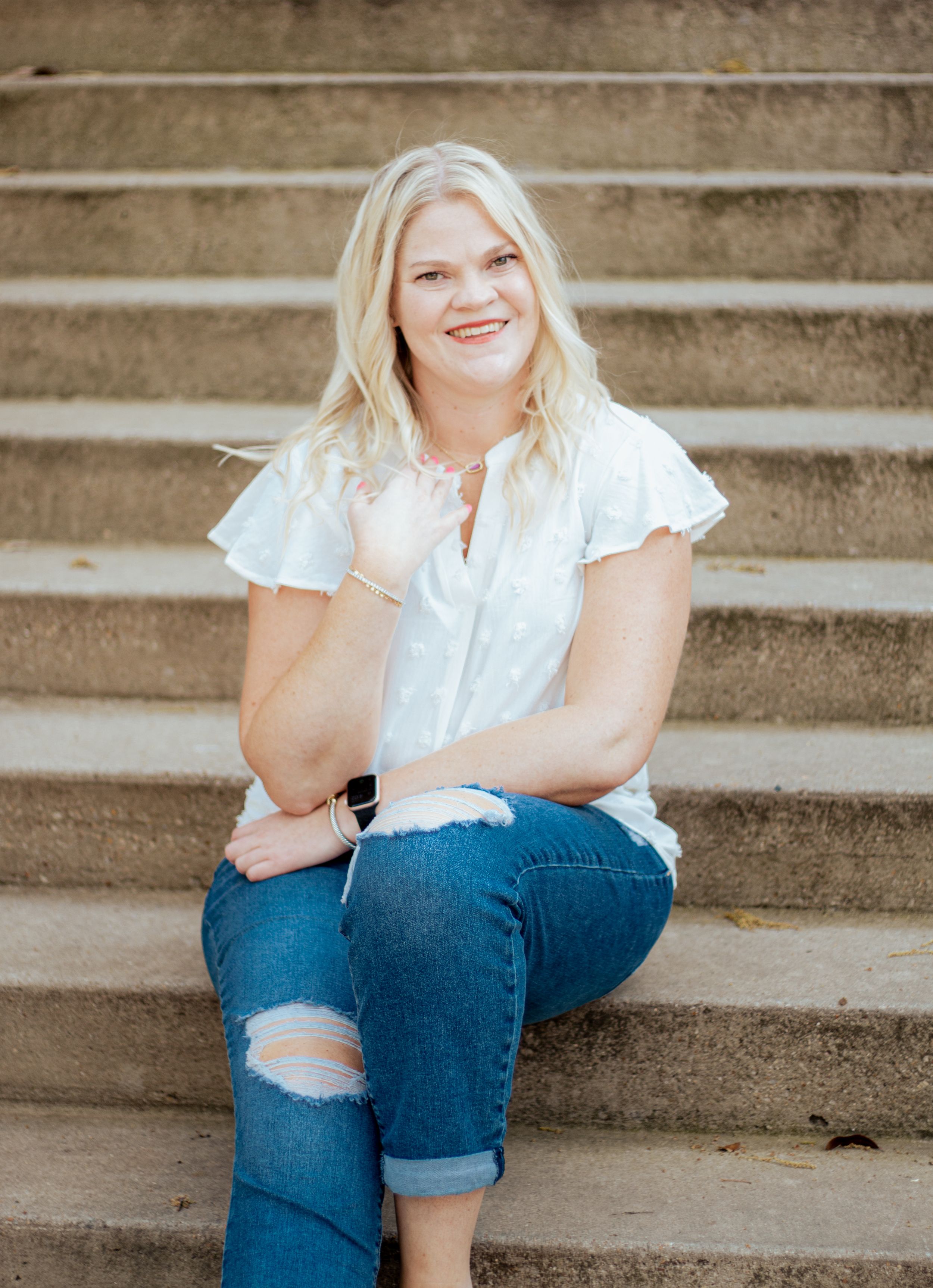 Person in white ruffled top and distressed jeans sits casually on concrete steps outdoors.