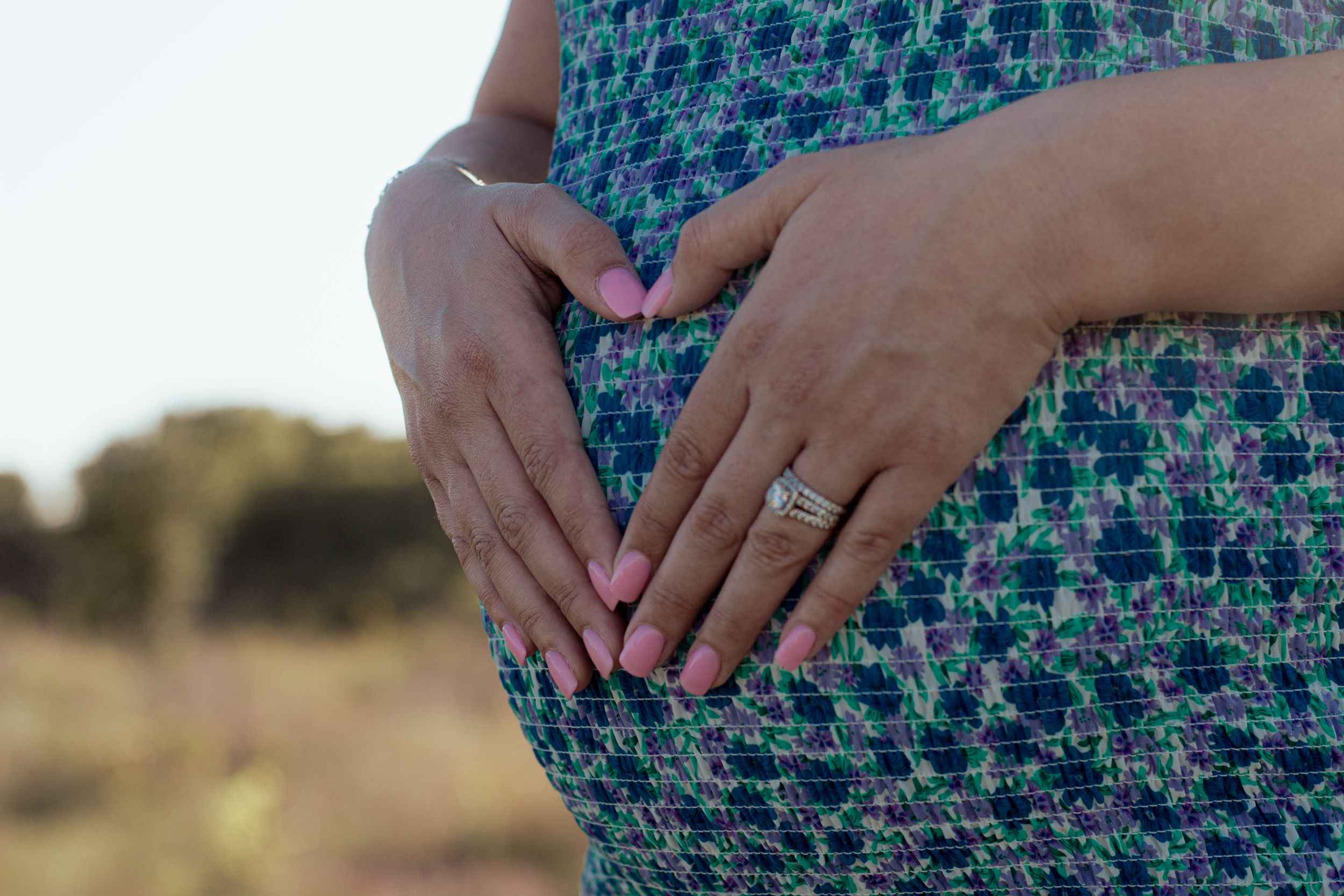 Hands wearing an engagement ring rest on a blue and green patterned dress during an outdoor photo session.