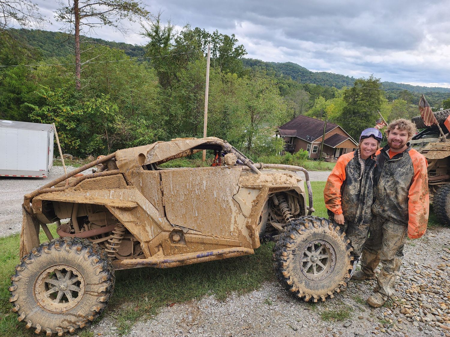 A mud-covered off-road vehicle sits beside two people in rain gear on a rural mountain trail.