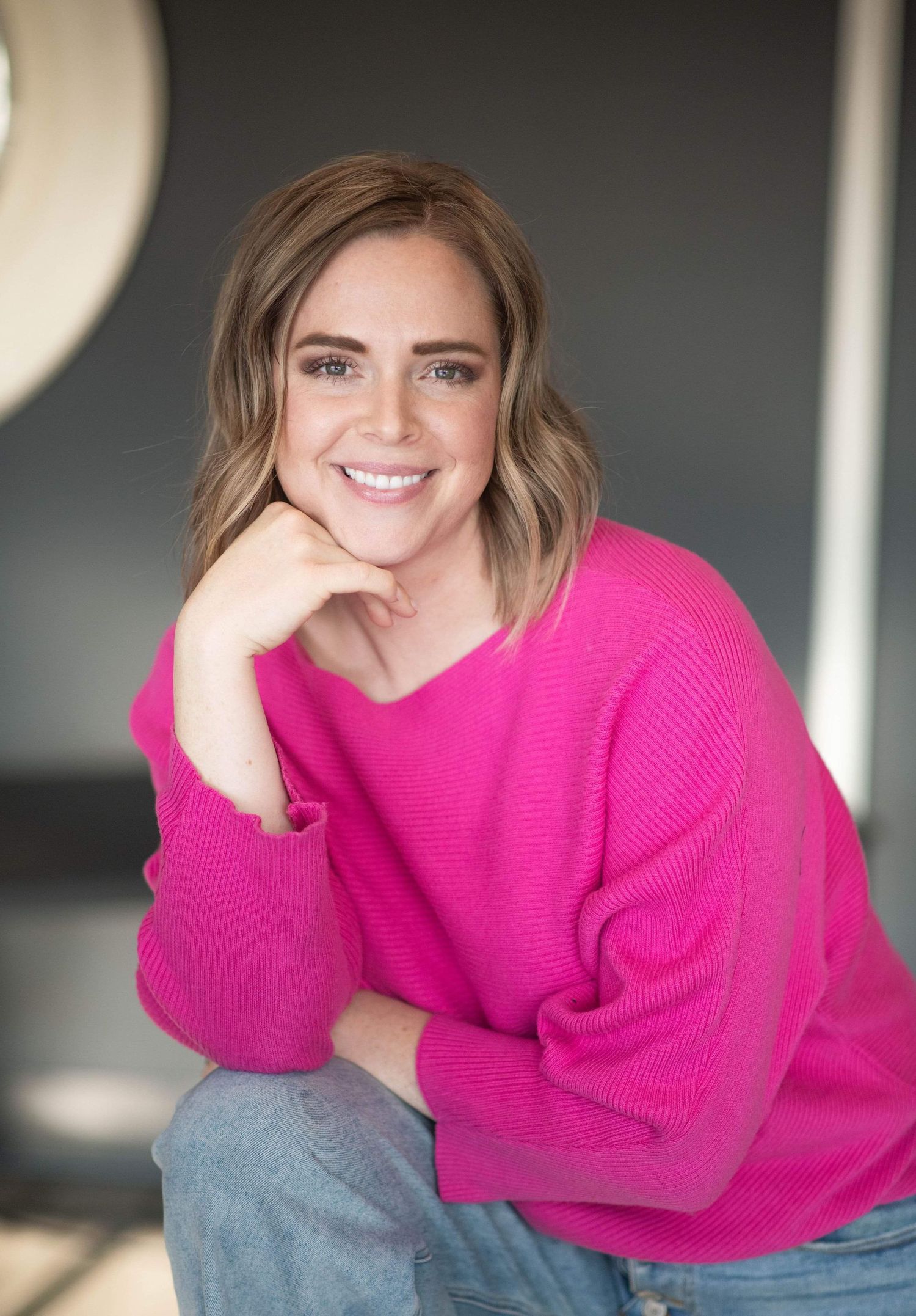 Smiling person wearing a bright pink sweater leans casually with hand on chin against light background.