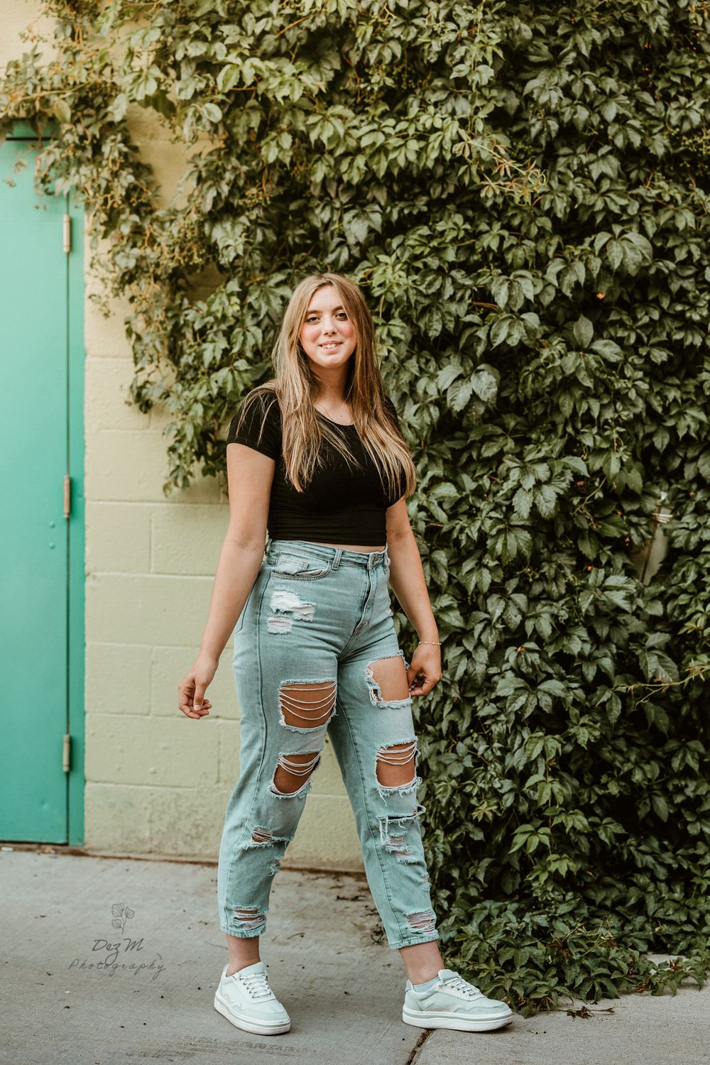 Person in distressed jeans poses casually against ivy-covered wall with turquoise door.