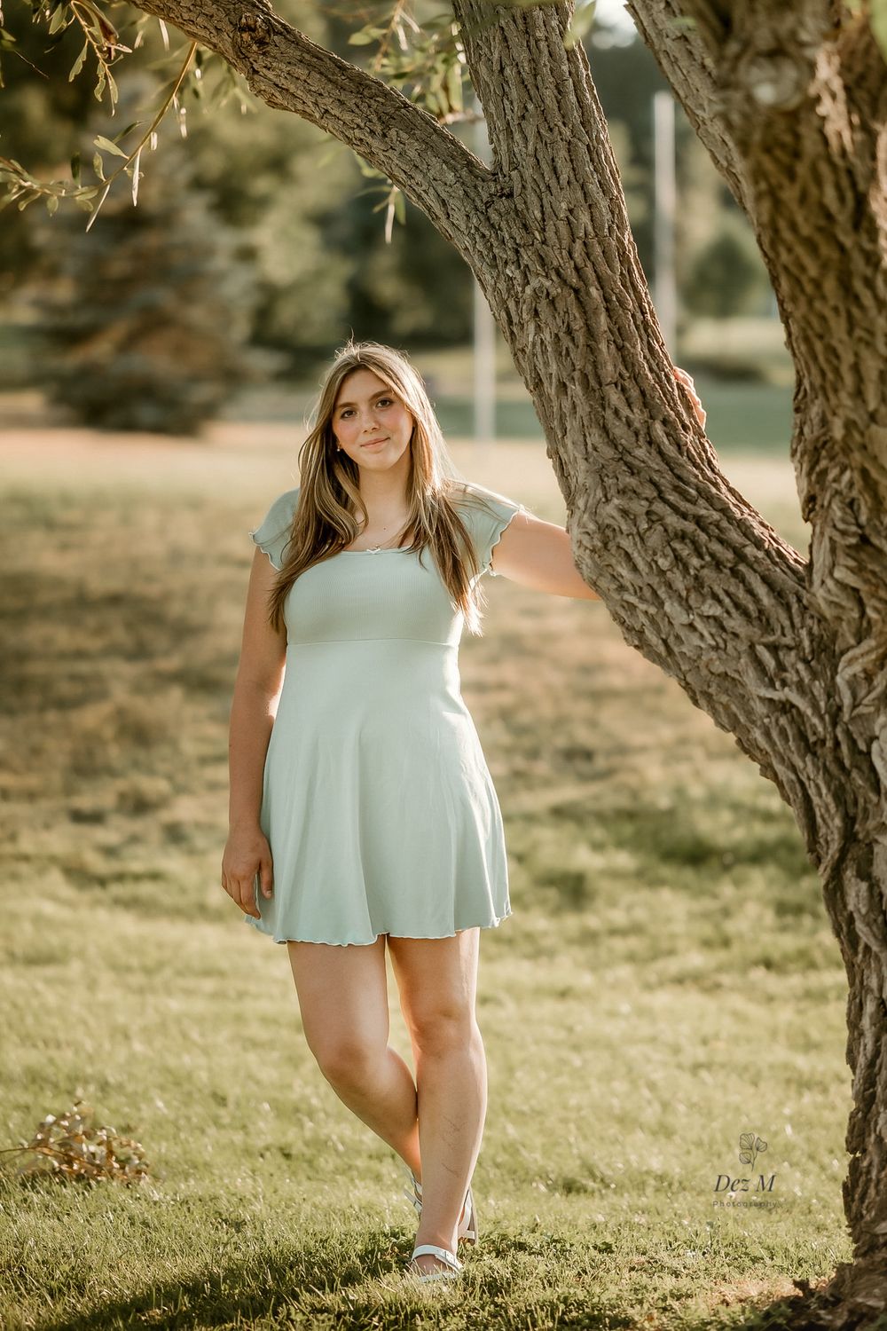 Person in white dress poses playfully under twisted tree branches in golden sunlight.