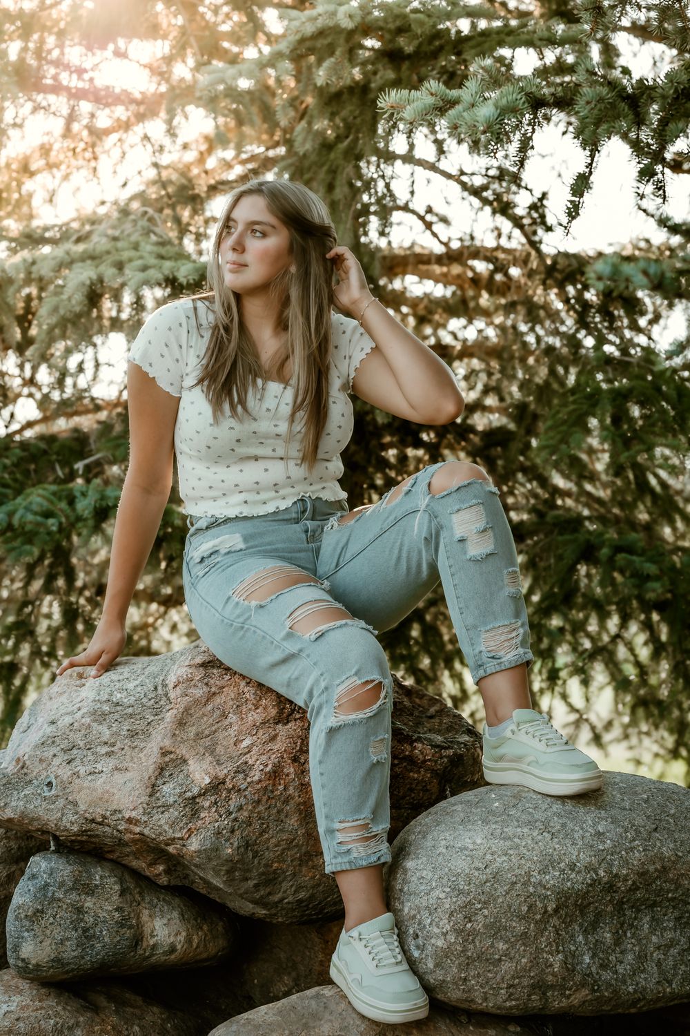 Person in ripped jeans and white shoes poses casually on large rocks at sunset.