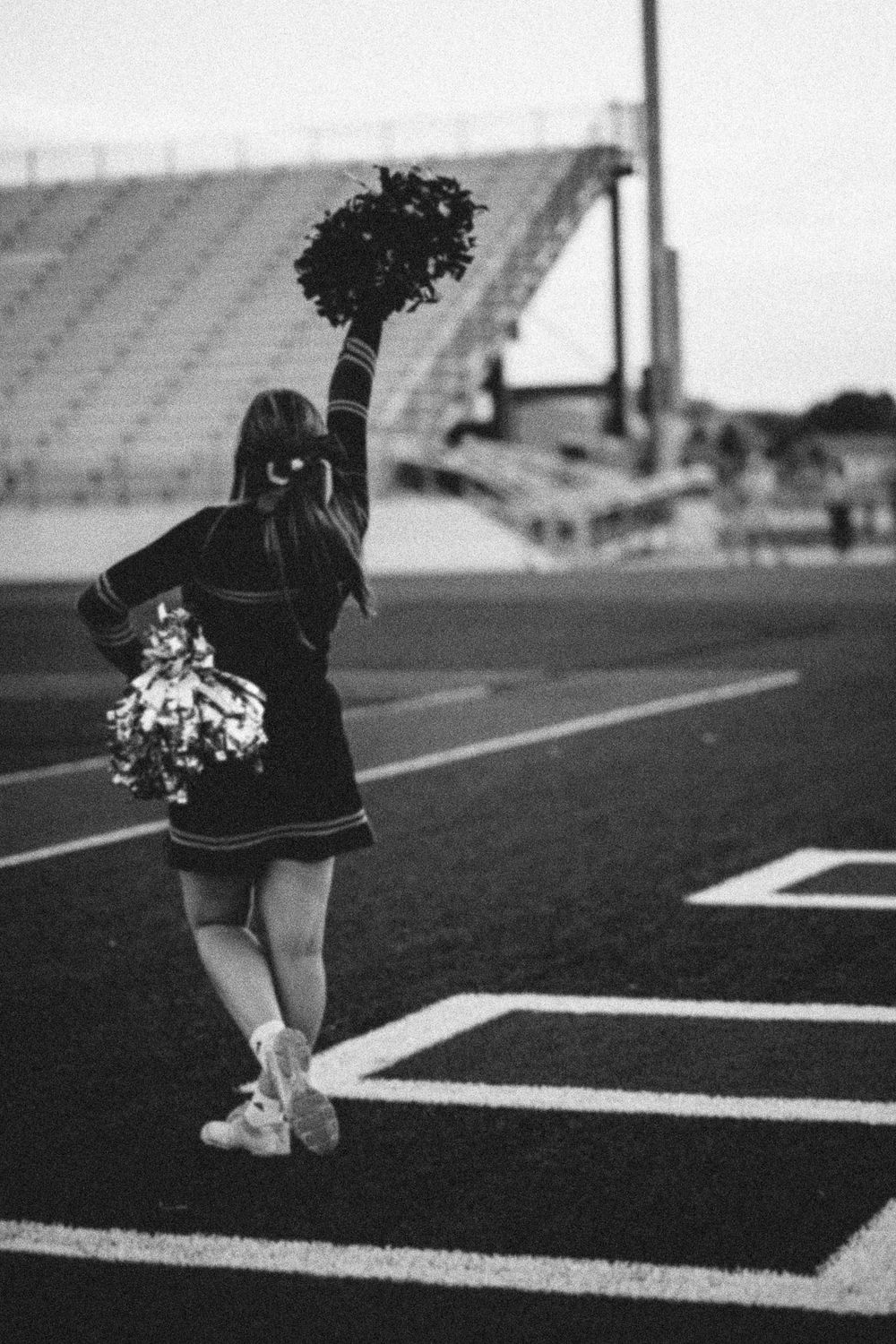 Black and white photo of cheerleader performing on football field with pom poms.