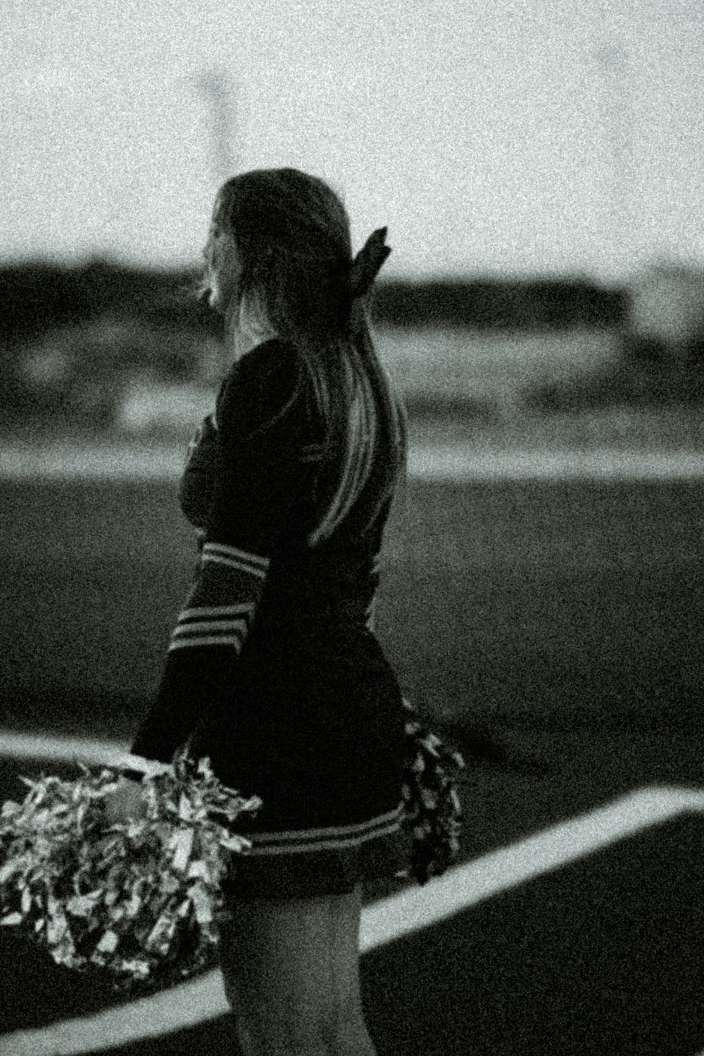 Black and white image of a cheerleader with pom poms against a dark background.