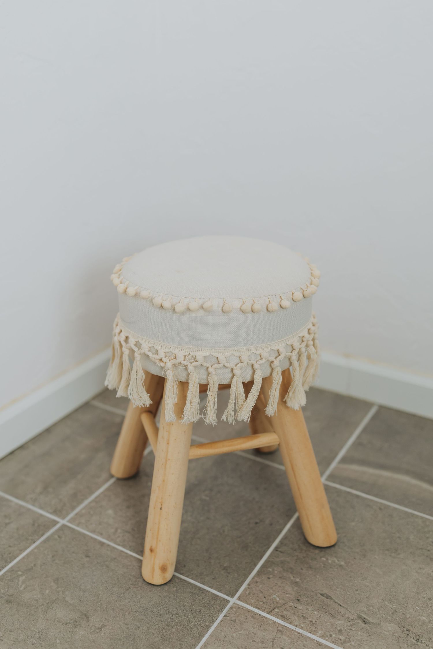 Round wooden stool with cream macrame trim displayed on gray tile flooring.