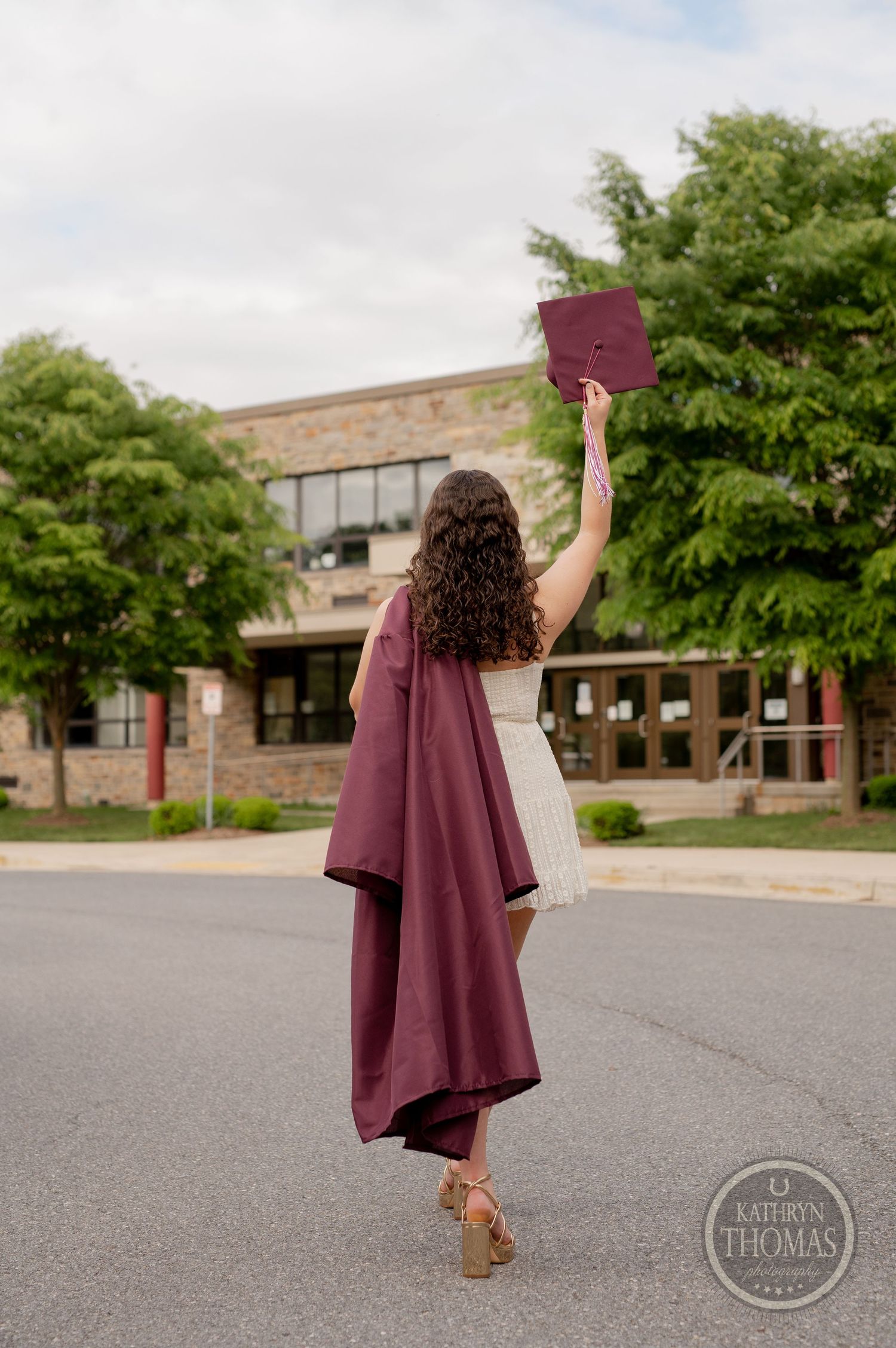 Graduate raises cap triumphantly while wearing burgundy gown and white dress outside school.