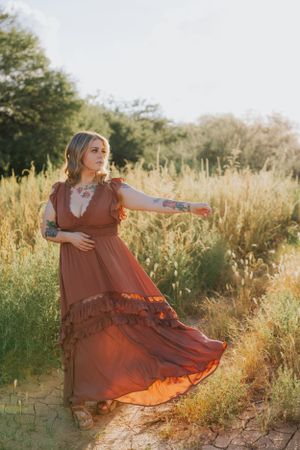 Woman in flowing rust-colored dress poses gracefully in golden field during sunset.