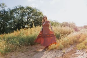 Ethereal outdoor portrait of woman in rust dress sitting among wild grasses at sunset.