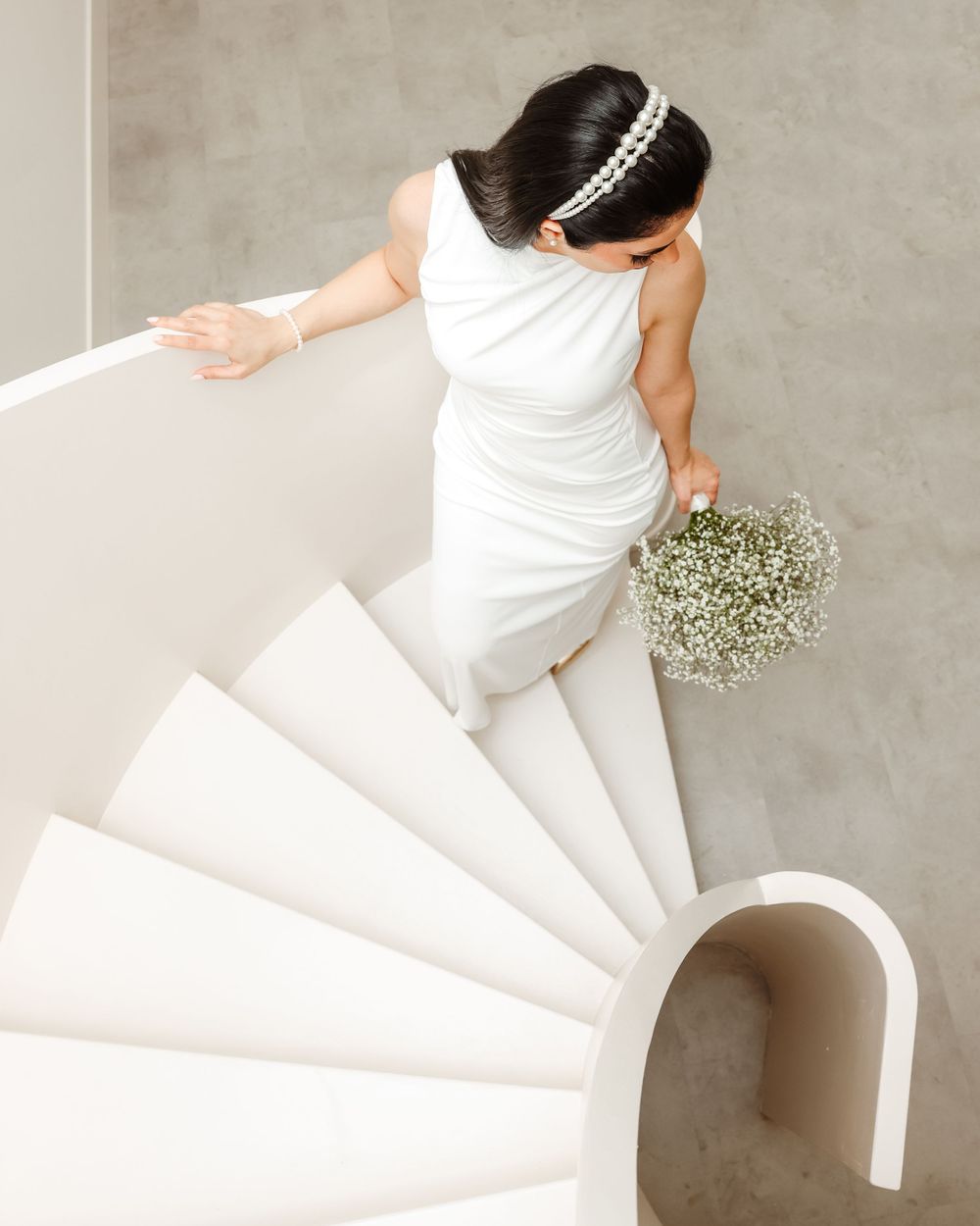 Aerial view of bride in white dress descending modern spiral staircase with baby's breath bouquet.