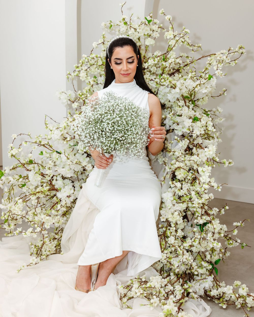 Bride in white dress poses with baby's breath floral arrangements against white backdrop.