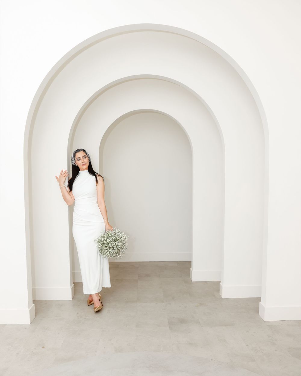 Elegant bride in white dress poses in minimalist arched hallway holding baby's breath bouquet.