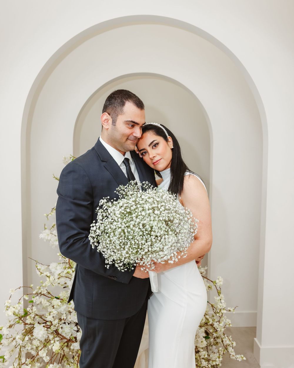 Stylish couple embraces in white archway surrounded by romantic baby's breath floral installation.