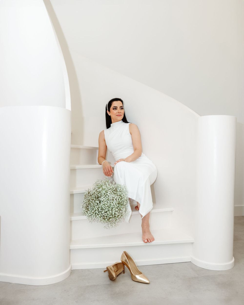 Bride in sleek white dress sits on modern staircase with baby's breath bouquet and gold heels.