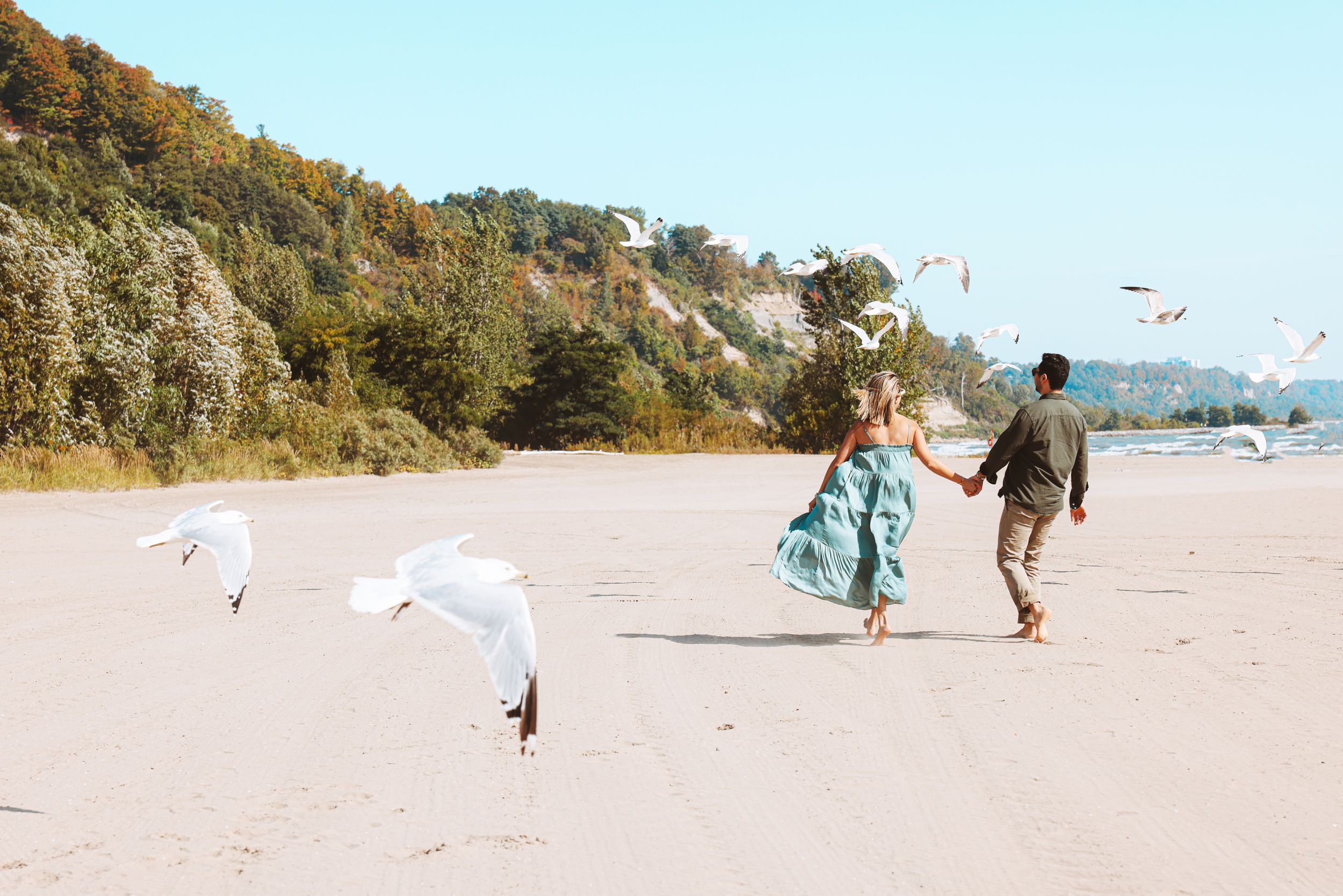 Two people run joyfully on beach while seagulls fly overhead against coastal cliffs.