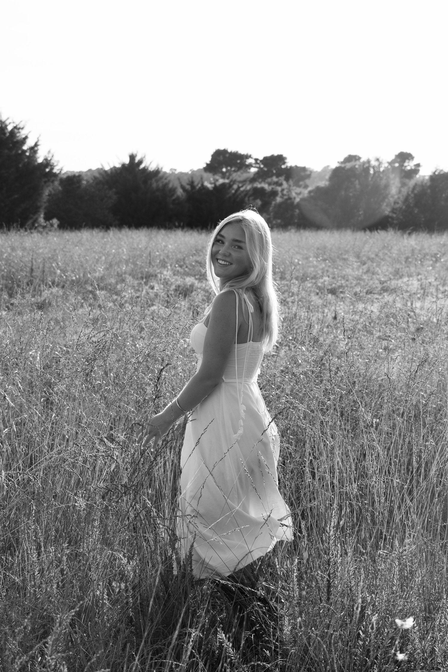 Black and white portrait of someone in a flowing white dress standing in a sunlit field of tall grass.
