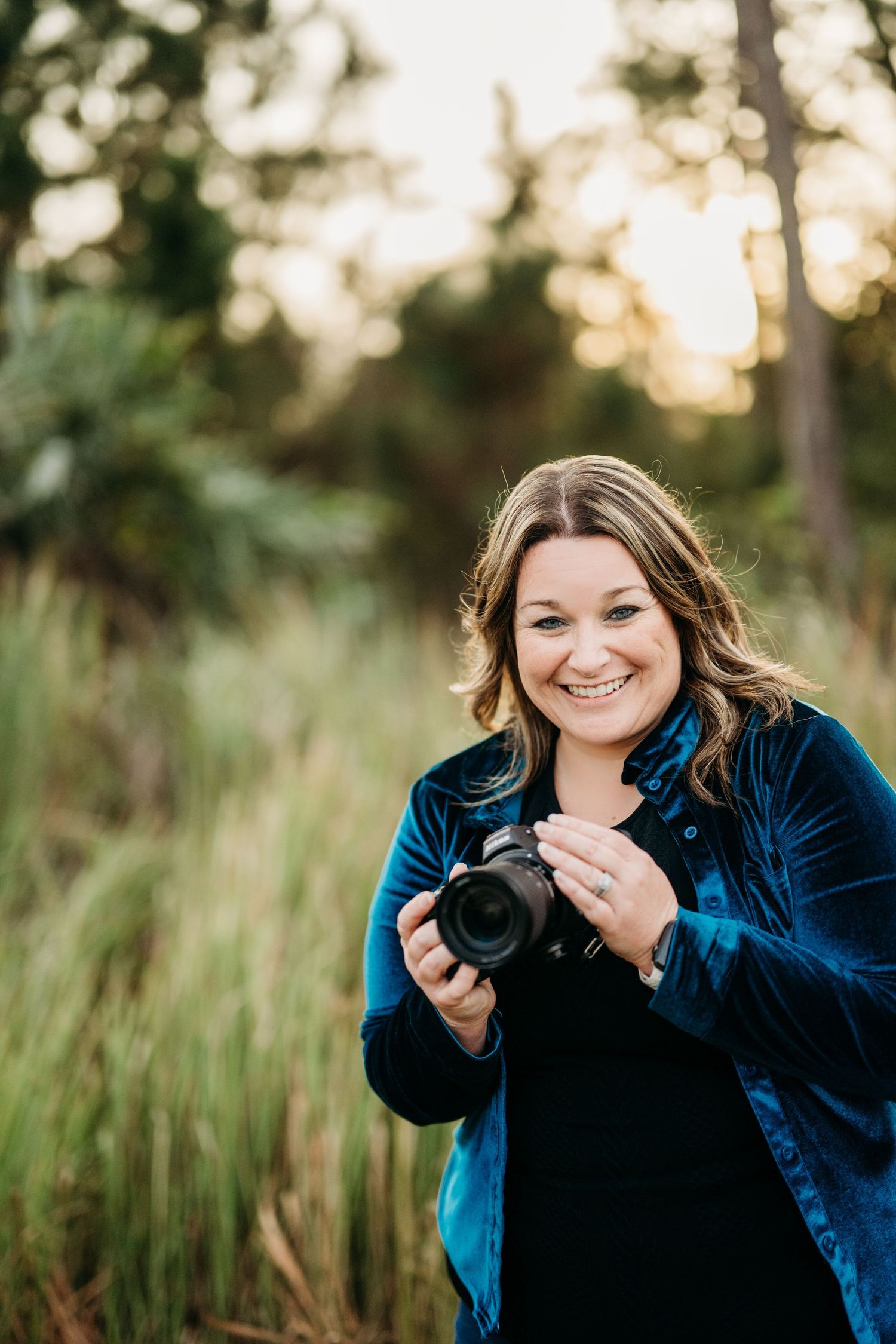 A photographer wearing a blue velvet jacket holds a camera while standing in tall grass at sunset.
