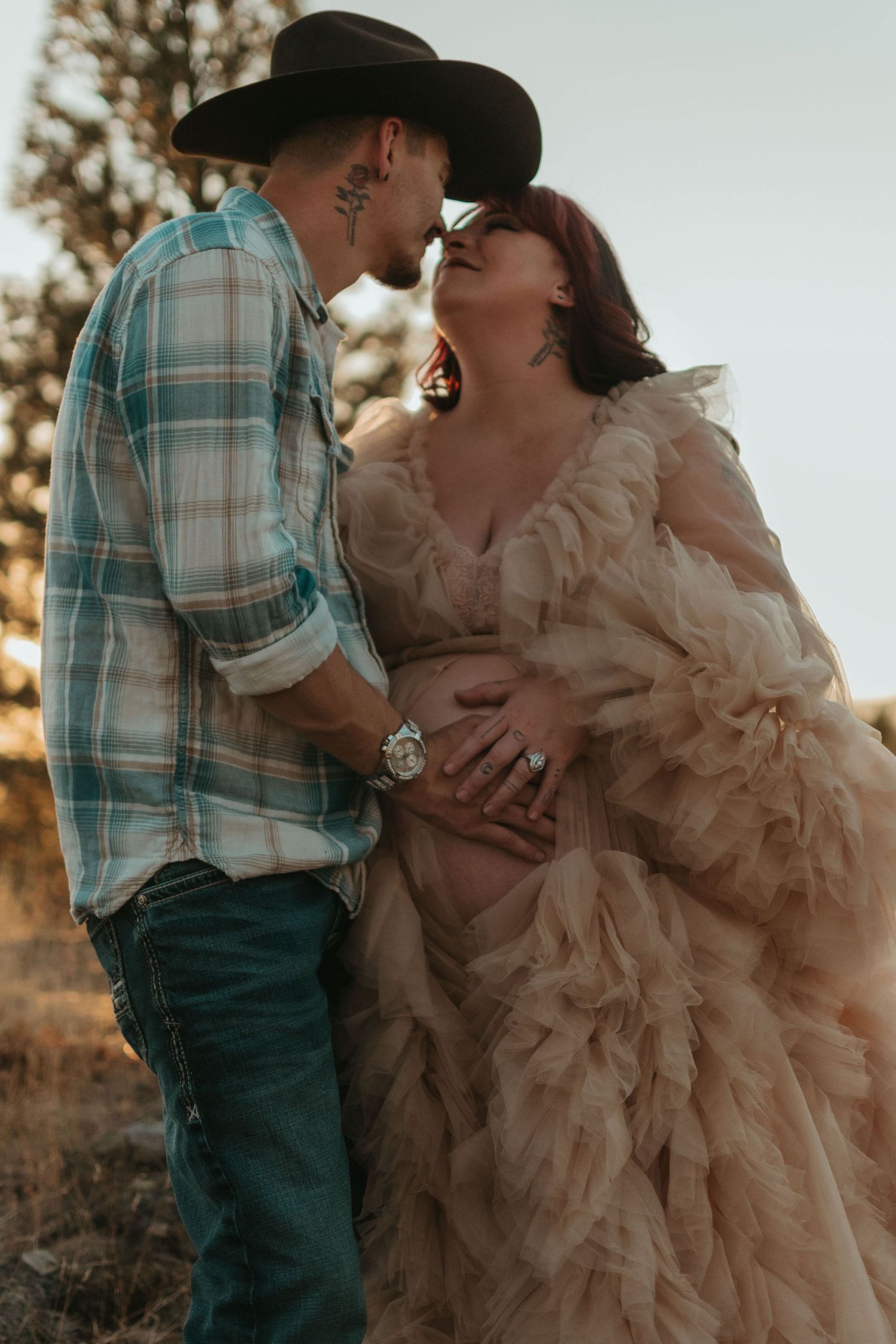 A romantic sunset maternity photo showing a couple in western attire and a flowing ruffled gown tenderly embracing outdoors.