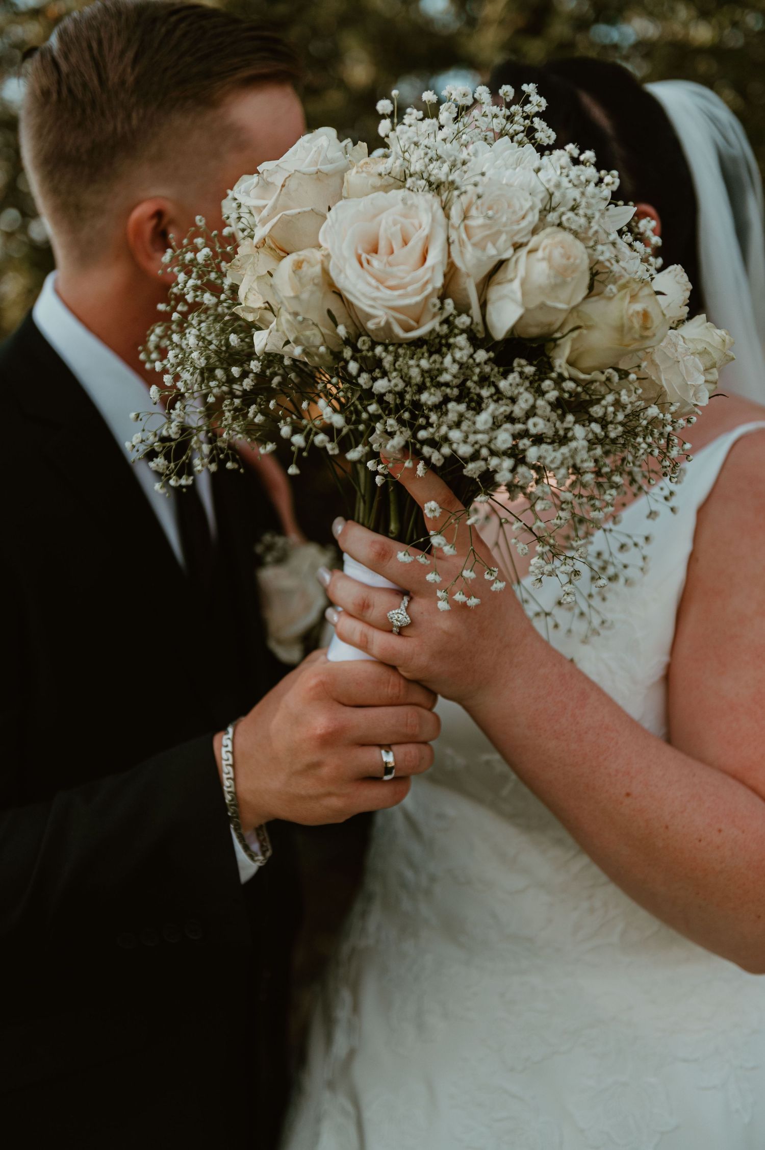 Close up of white rose and baby's breath wedding bouquet with hands showing rings.