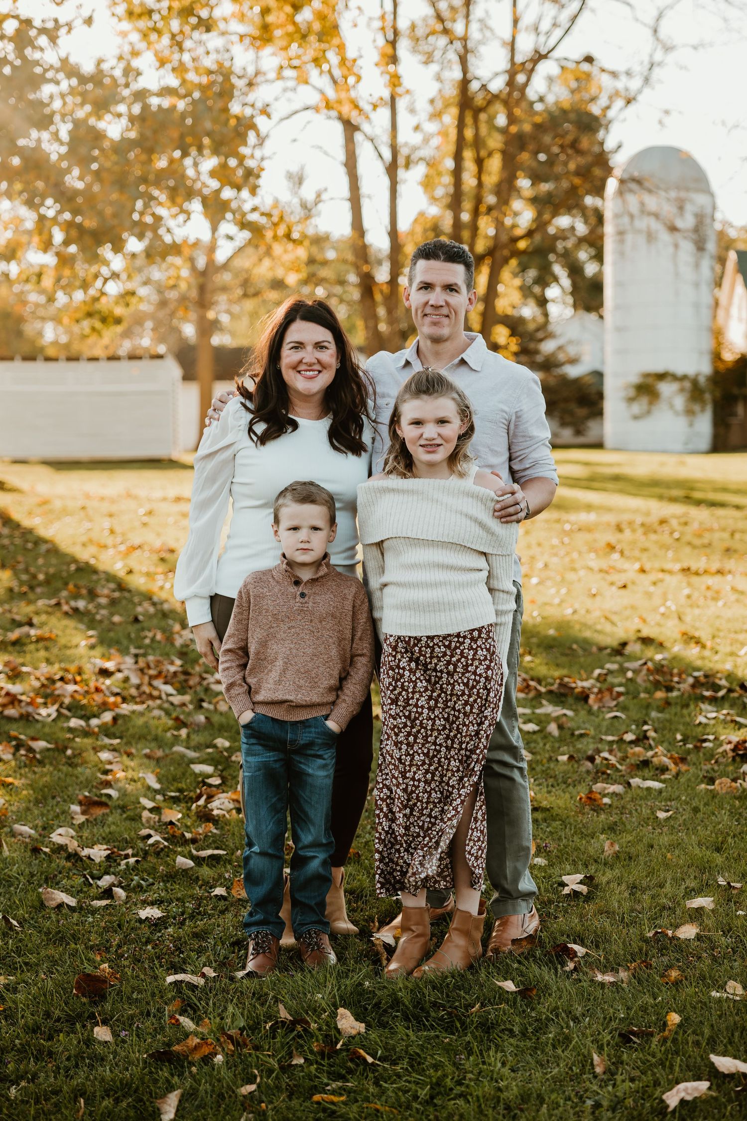 Family of four poses together with autumn trees and white building in background. Michigan Detroit Novi Northville Portrait Photographer