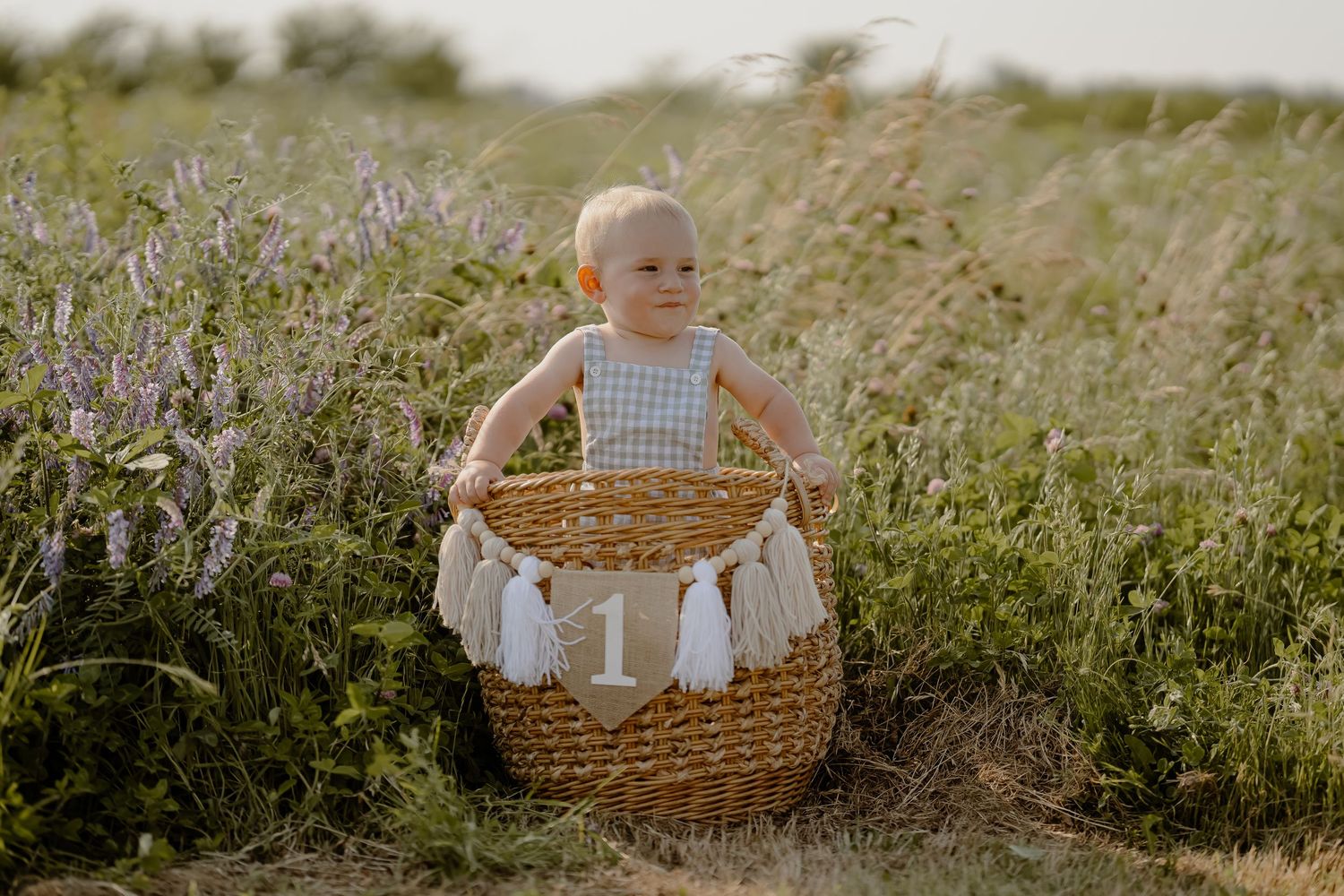 A baby in tan overalls crawls on a white blanket in a sunny park setting with a blue milestone marker.