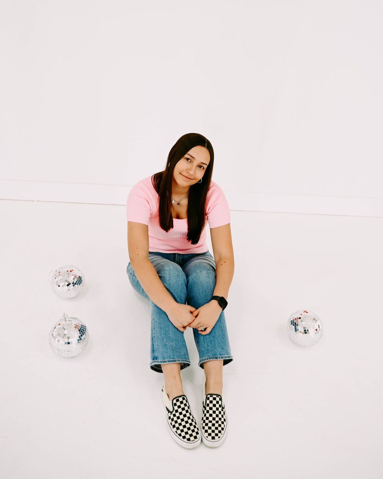 Person wearing checkerboard slip-on shoes and jeans sits on white floor with disco balls in studio setting.