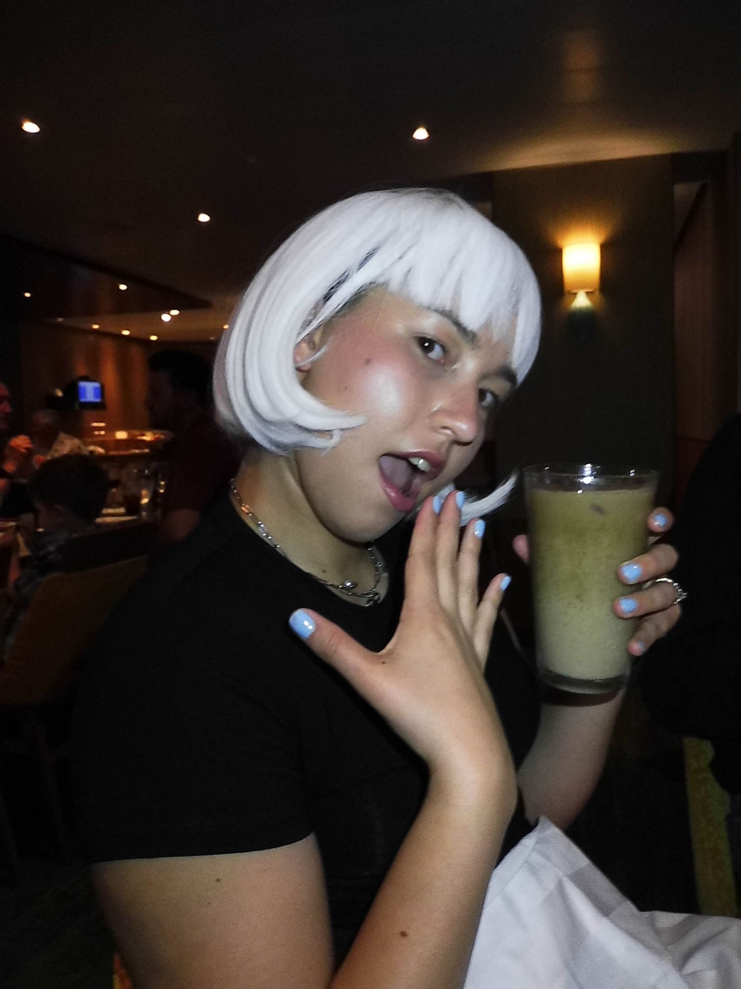 A person with white hair sips a green smoothie drink in a dimly lit room.