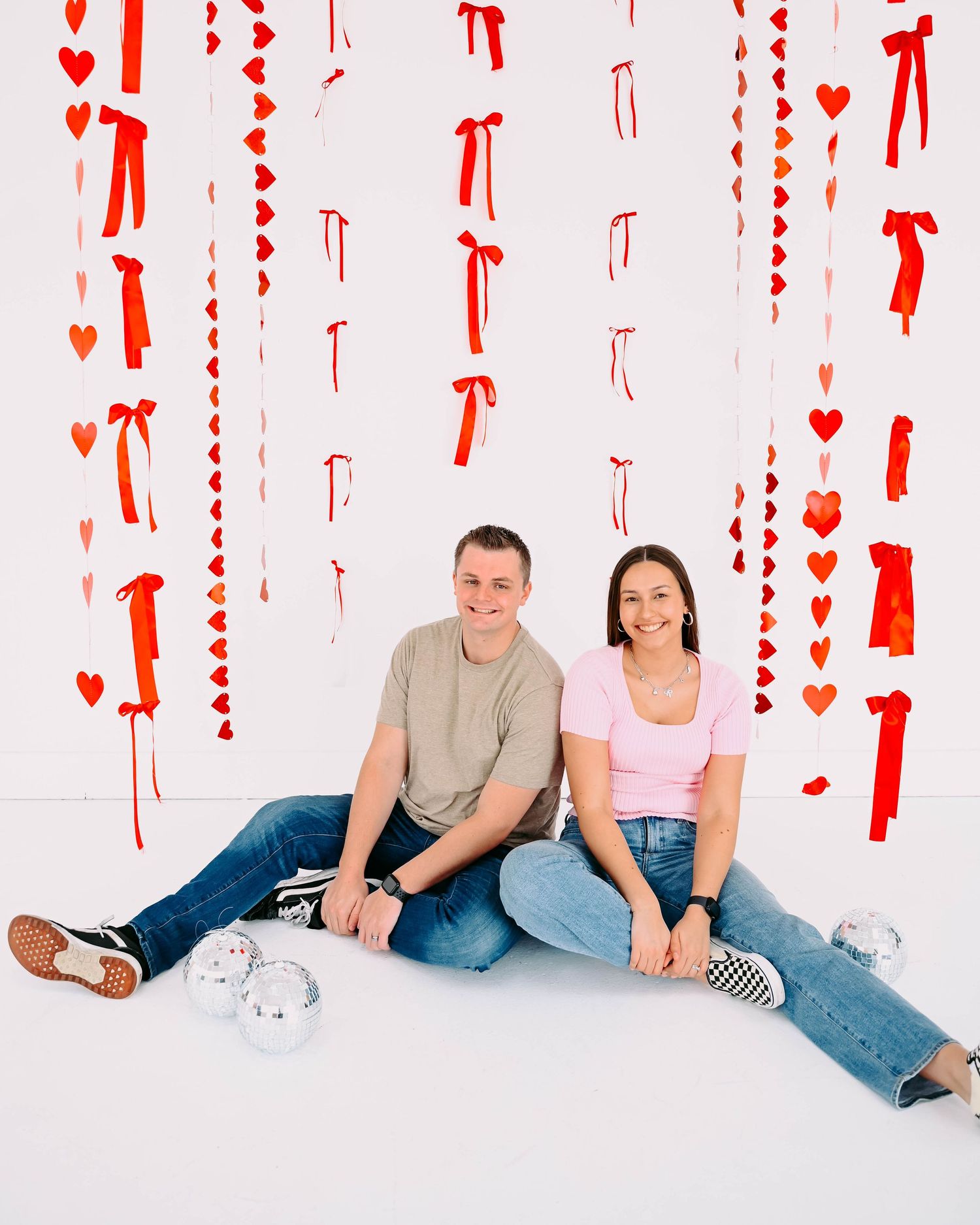 Two people sitting on white floor with red heart and ribbon decorations hanging on wall behind them.