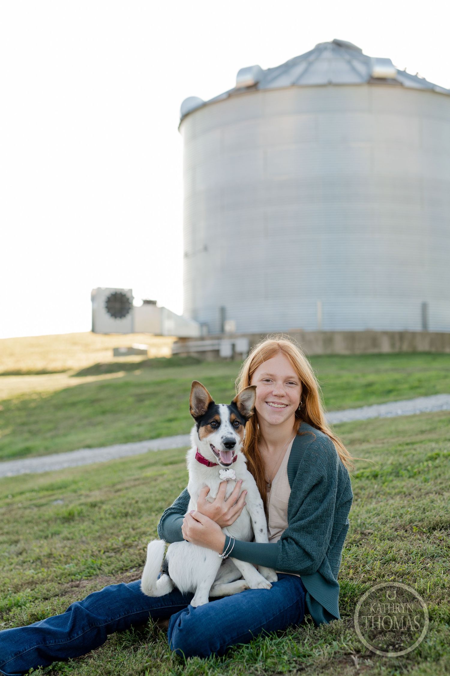 A sweet black and white spotted dog sits with a person in front of a large grain silo on a sunny day.