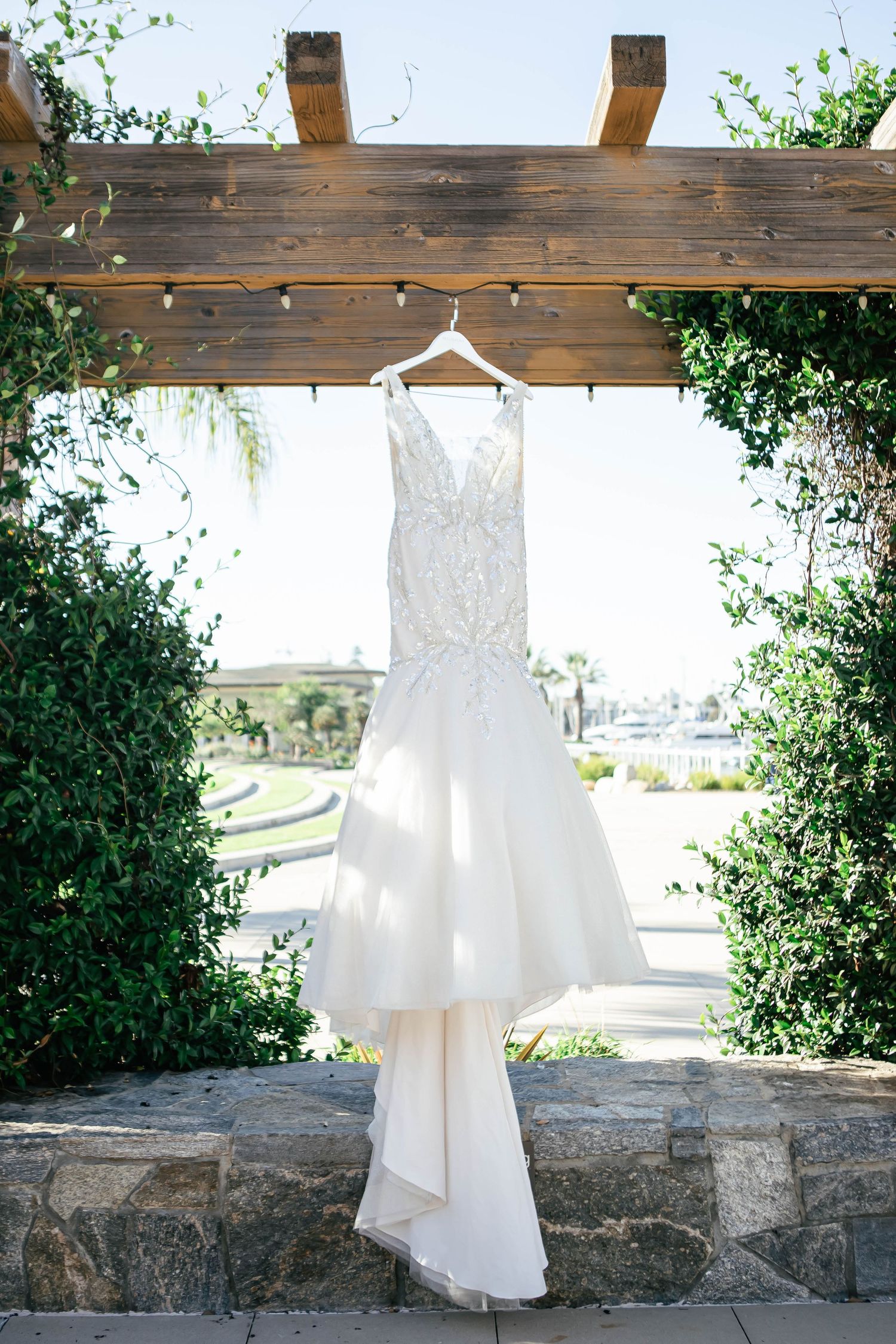 White wedding dress hanging on wooden pergola with stone wall and greenery.