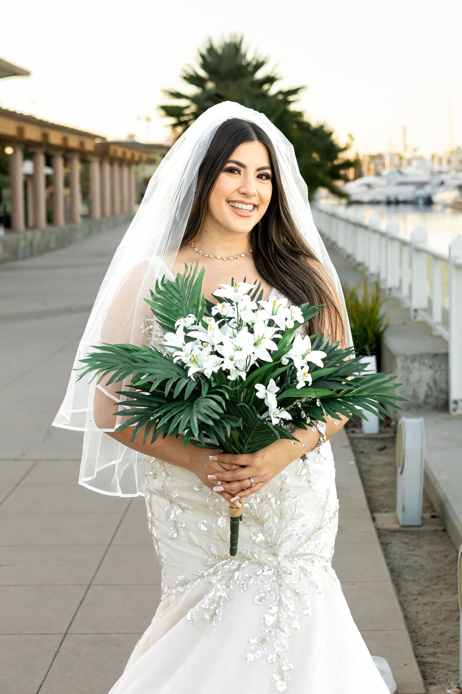 Bride in white lace dress holding tropical bouquet with white flowers and palm leaves.