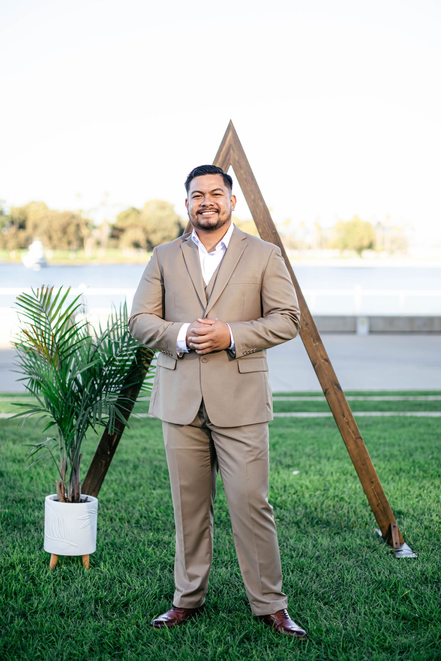 Person in tan suit poses with wooden triangle arch and potted palm on grass.
