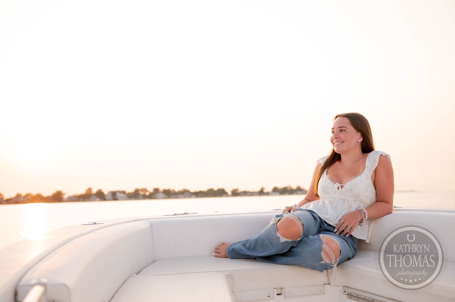 Woman lounges on white boat cushions against serene waterfront sunset view.