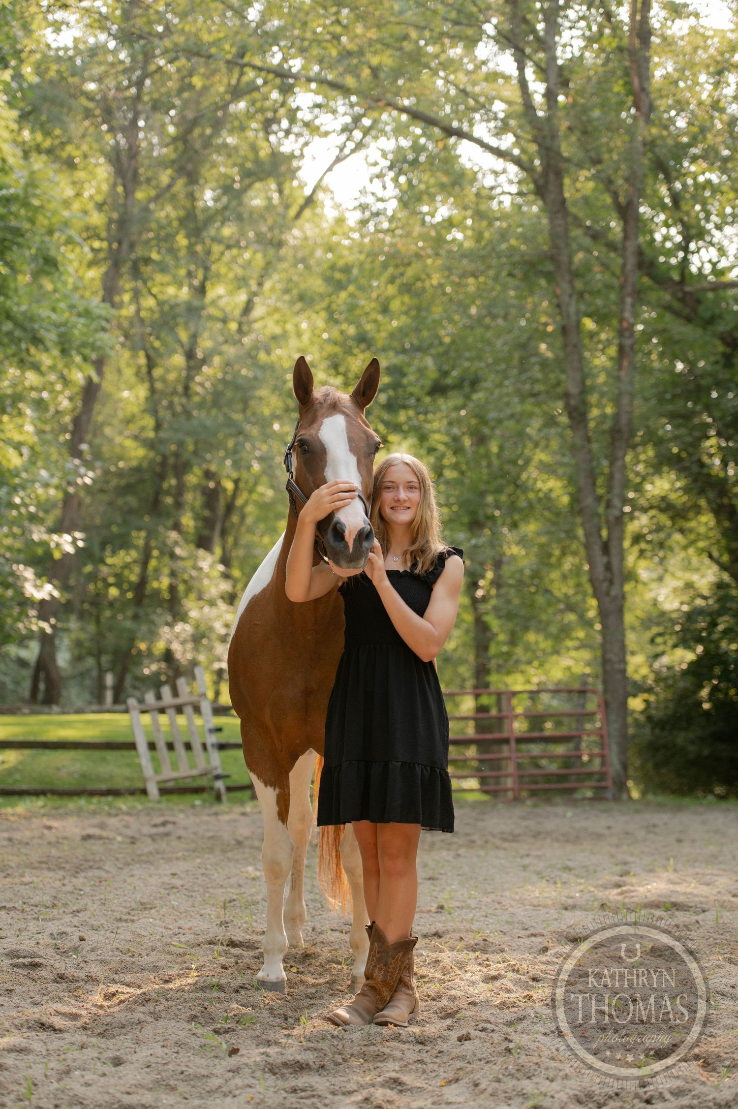 A person in boots and a black dress stands next to their paint horse in a fenced outdoor area.