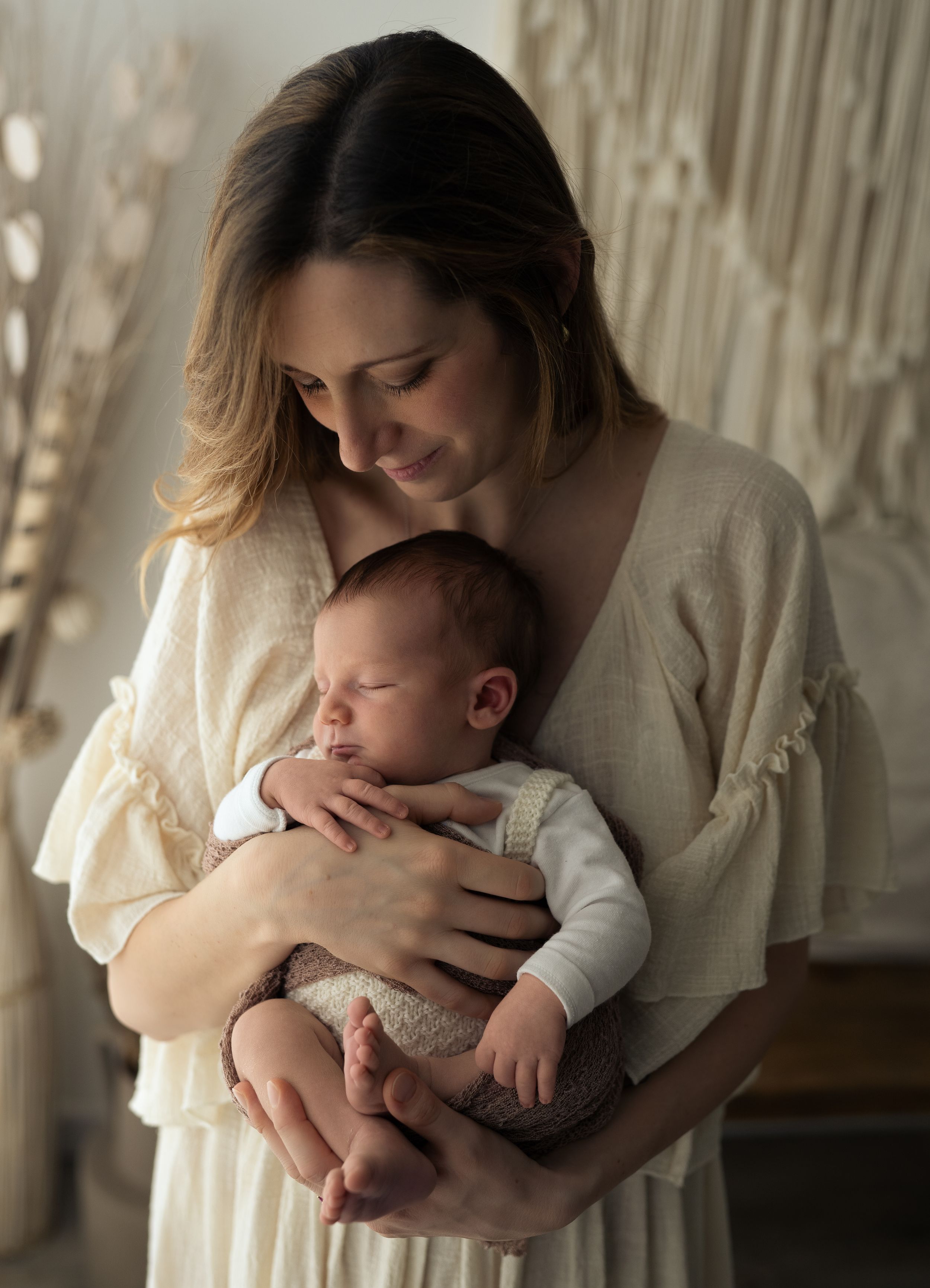 Neo mamma che tiene in braccio il suo bimbo di poche settimane ed entrambi indossano abiti dai toni neutri e romantici