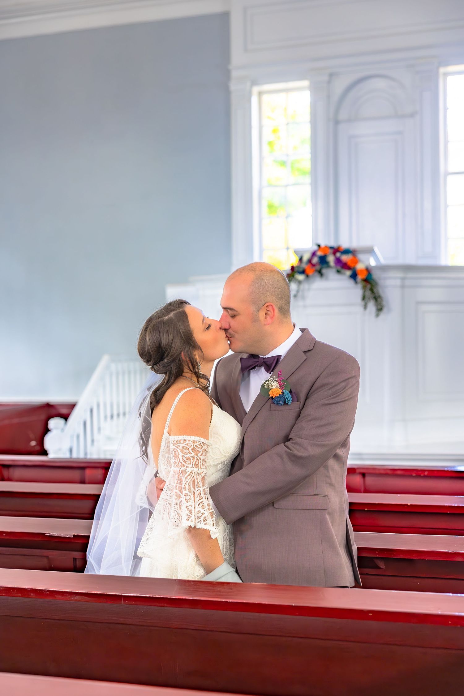 A couple shares a romantic kiss while sitting in red church pews during their wedding ceremony.
