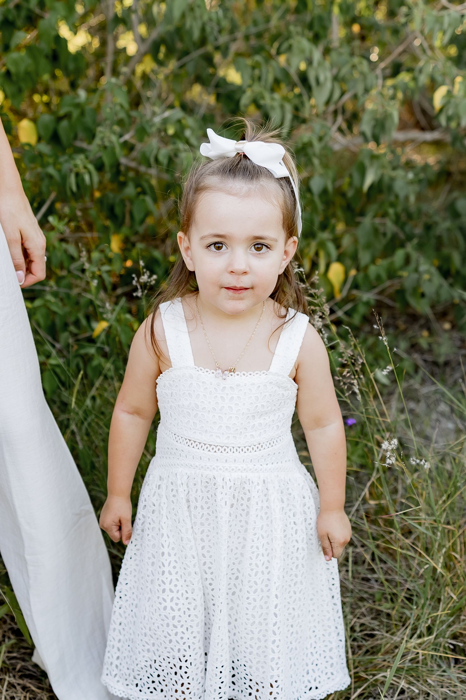 A young child in a white eyelet lace dress and bow stands outdoors against a natural green background.