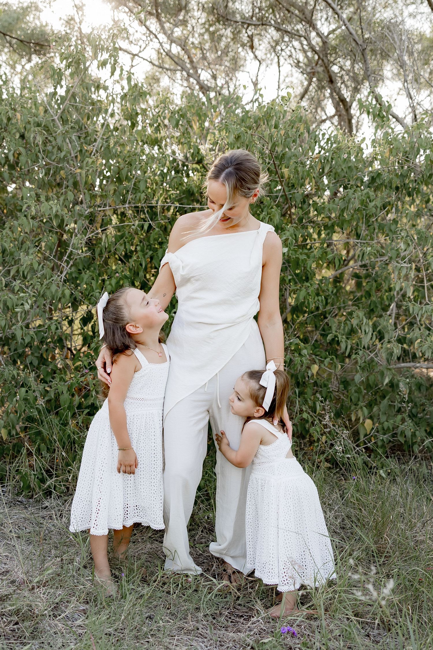 A sweet family moment captured in white outfits against a natural outdoor backdrop with greenery.
