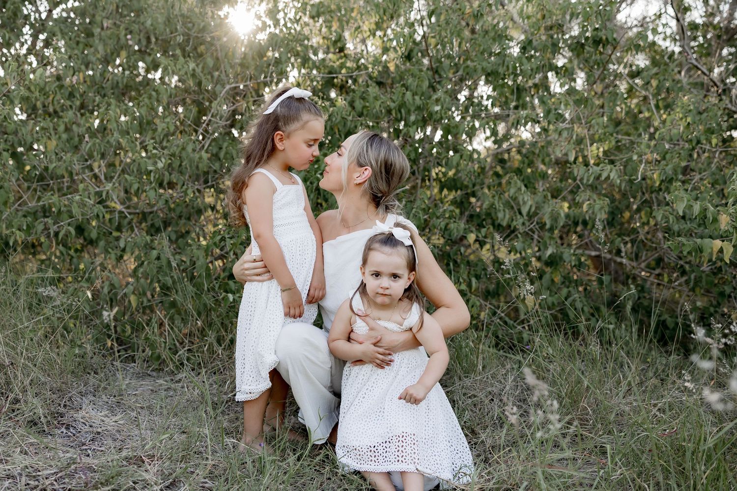 A family dressed in all white embraces during a golden hour photoshoot in a natural outdoor setting.