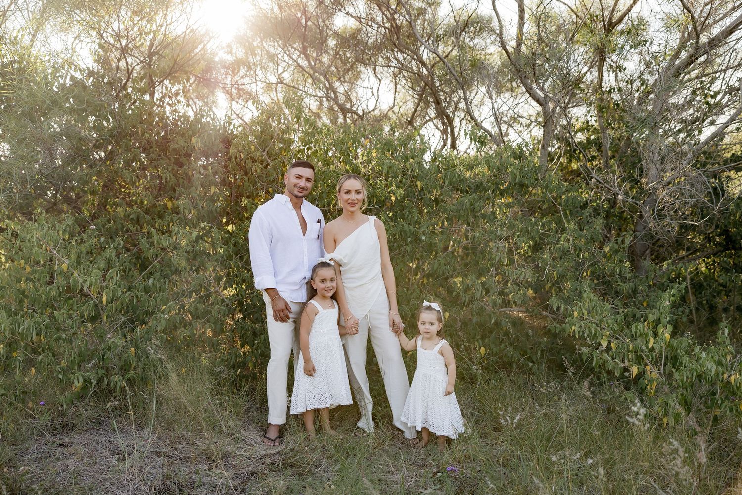 A family dressed in white outfits poses together in a natural outdoor setting at sunset.