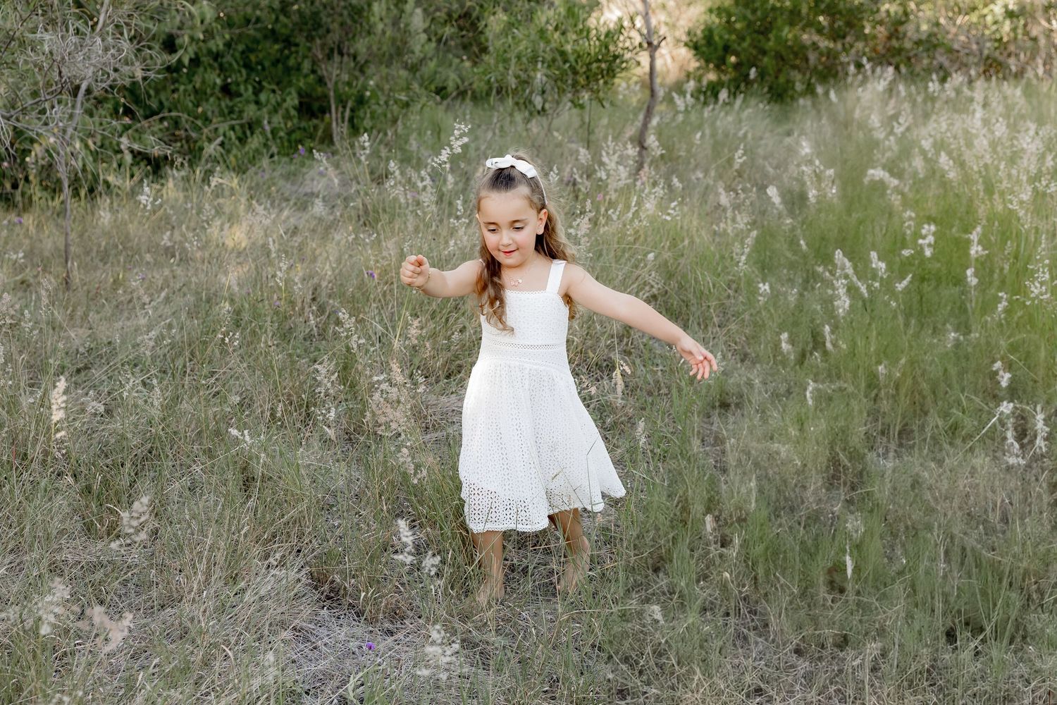 Family wearing white outfits twirls and plays together in a grassy meadow during golden hour.