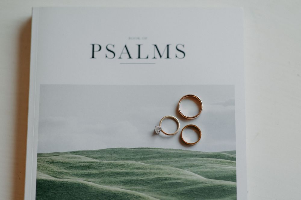 Three gold wedding rings rest on a book titled Psalms with a serene green landscape design on the cover.
