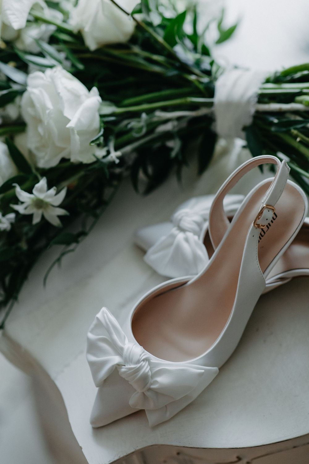 Elegant white bridal shoes with bow detail alongside white roses and greenery.