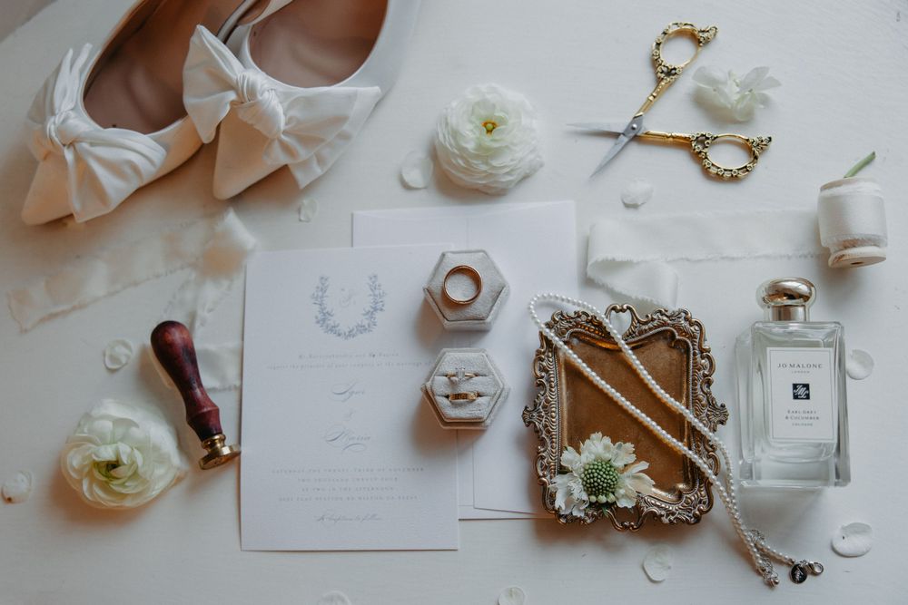 Wedding accessories flatlay with white shoes, perfume bottle, jewelry, flowers and invitation on light background.