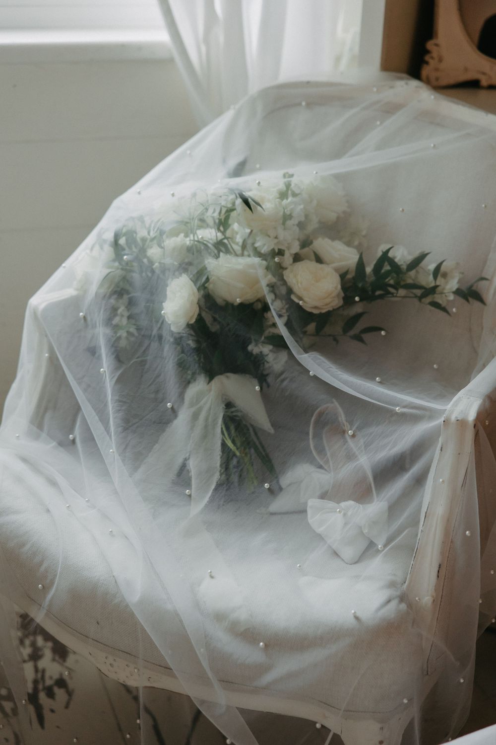 White roses and greenery bouquet draped with ethereal tulle veil in natural window light.