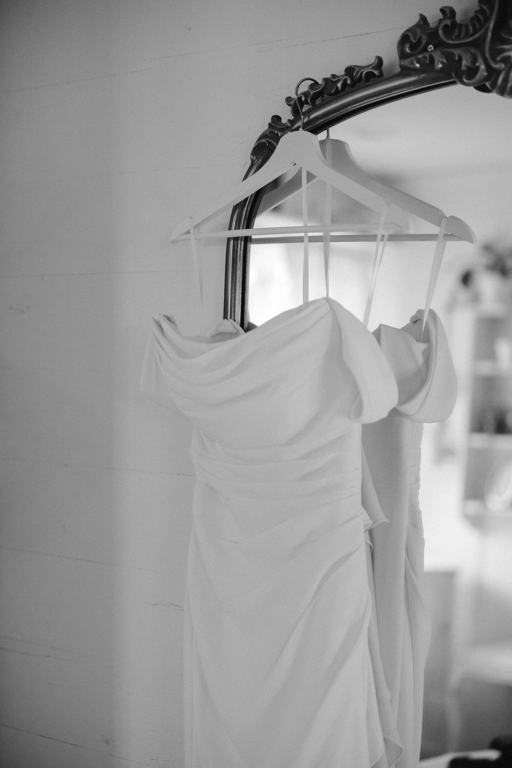 A white wedding dress hangs on a transparent hanger against an ornate mirror in black and white.